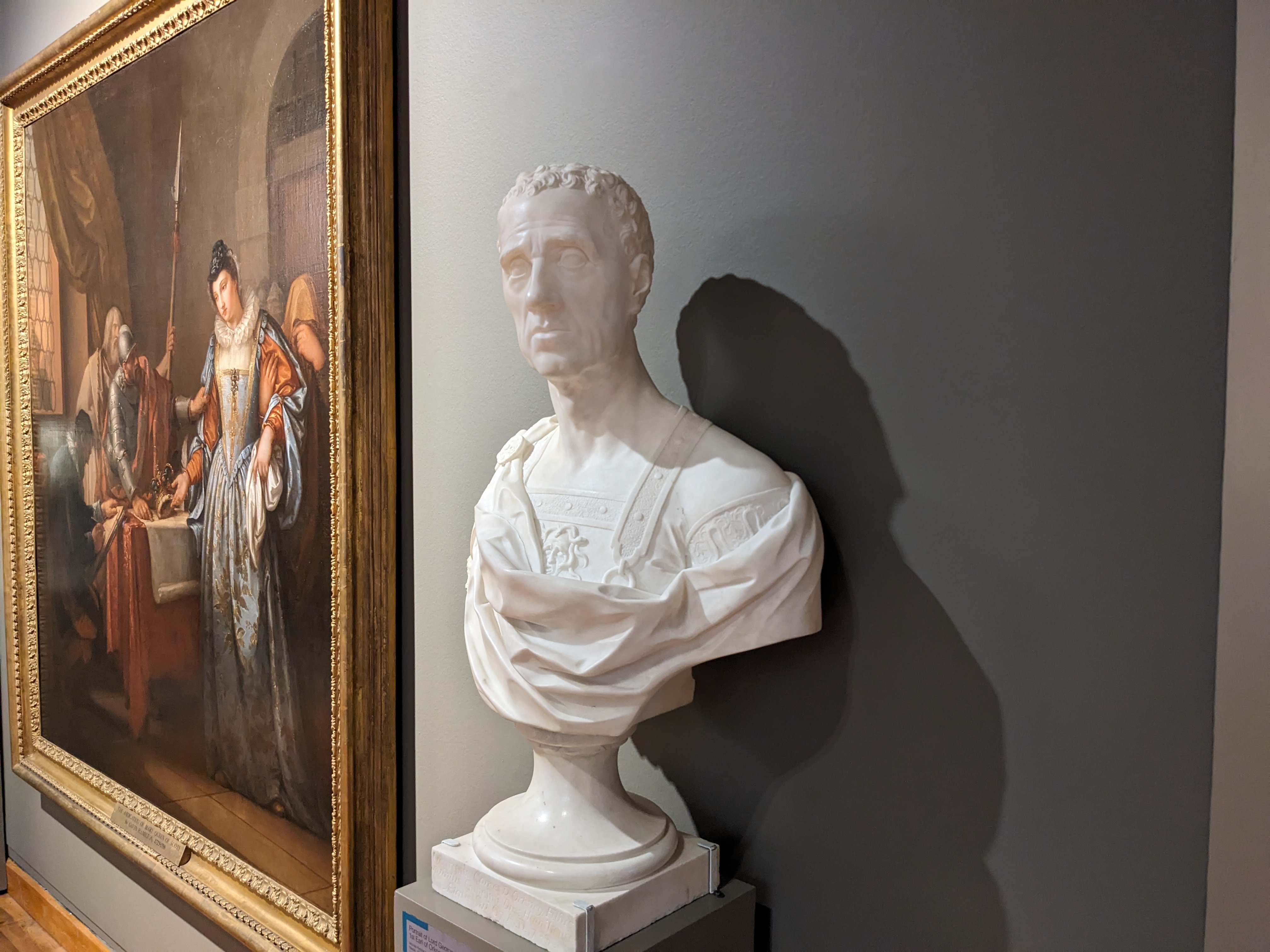 White marble bust of a man dressed in classical Roman attire. The sculpture is set against a grey background. A large painting is hung on the wall to the left of the sculpture.