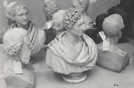 Black and white photograph showing a collection of classical marble busts arranged closely together on a surface. Busts feature detailed hair and clothing textures, with some having tags attached, indicating they may be part of a museum collection.
